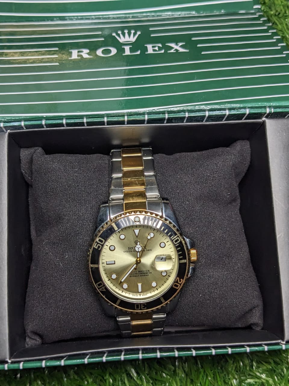 Gold and silver watch in a Rolex box on a grassy surface